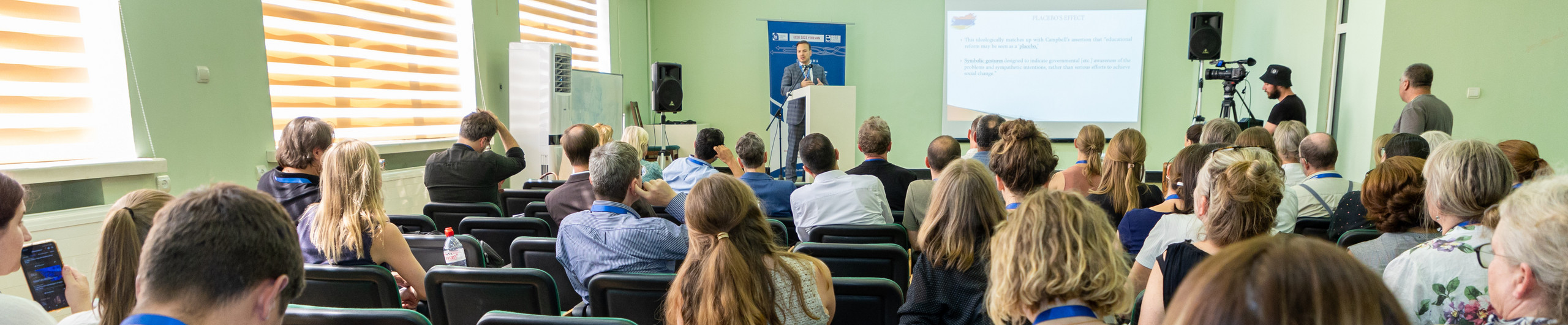 Audience from the back with one speaker and screen showing a presentation.